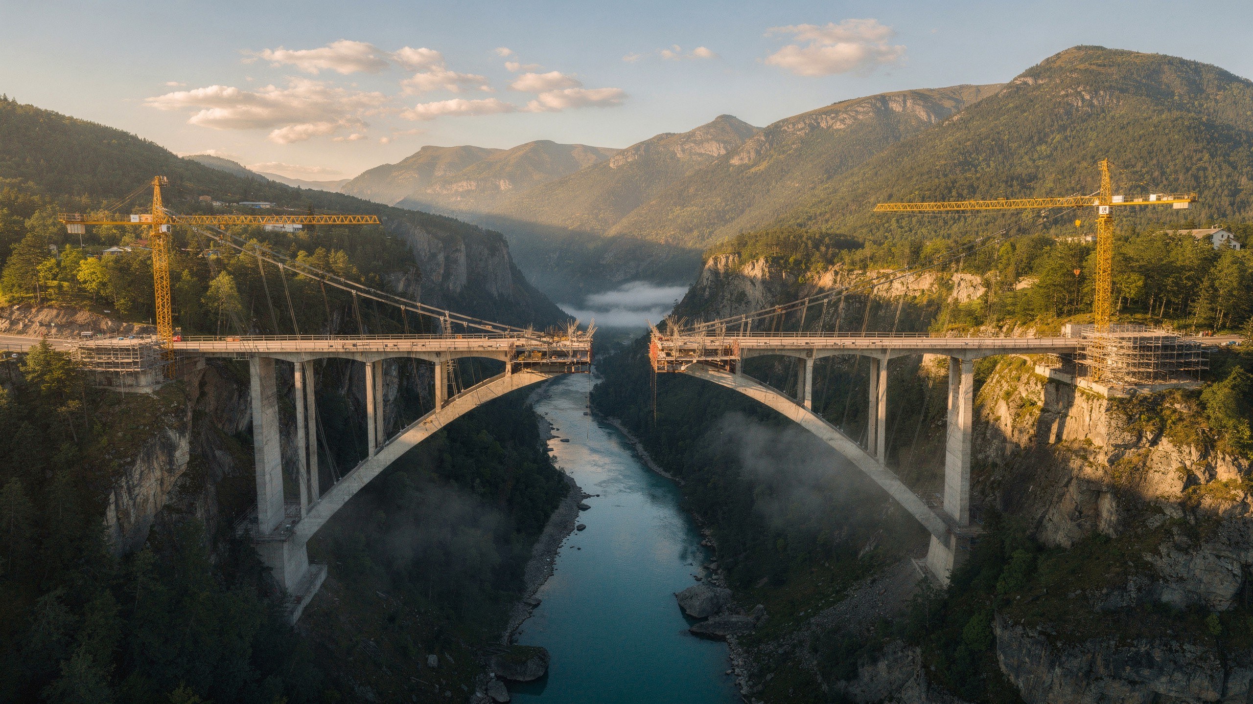Two bridge sections reach toward each other across a river gorge in warm light.